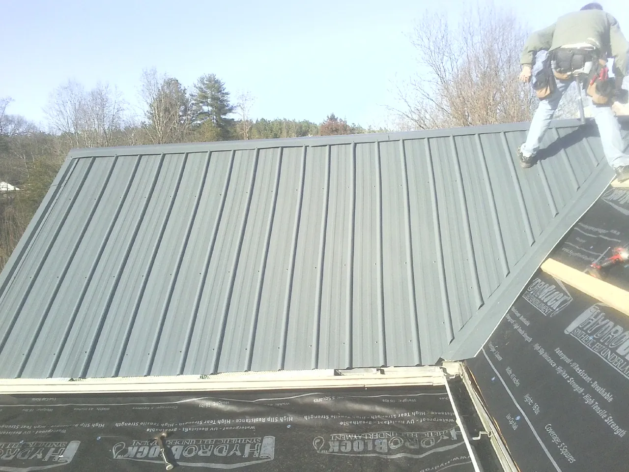 Crew installing dark green standing seam panels over synthetic underlayment in Low Moor