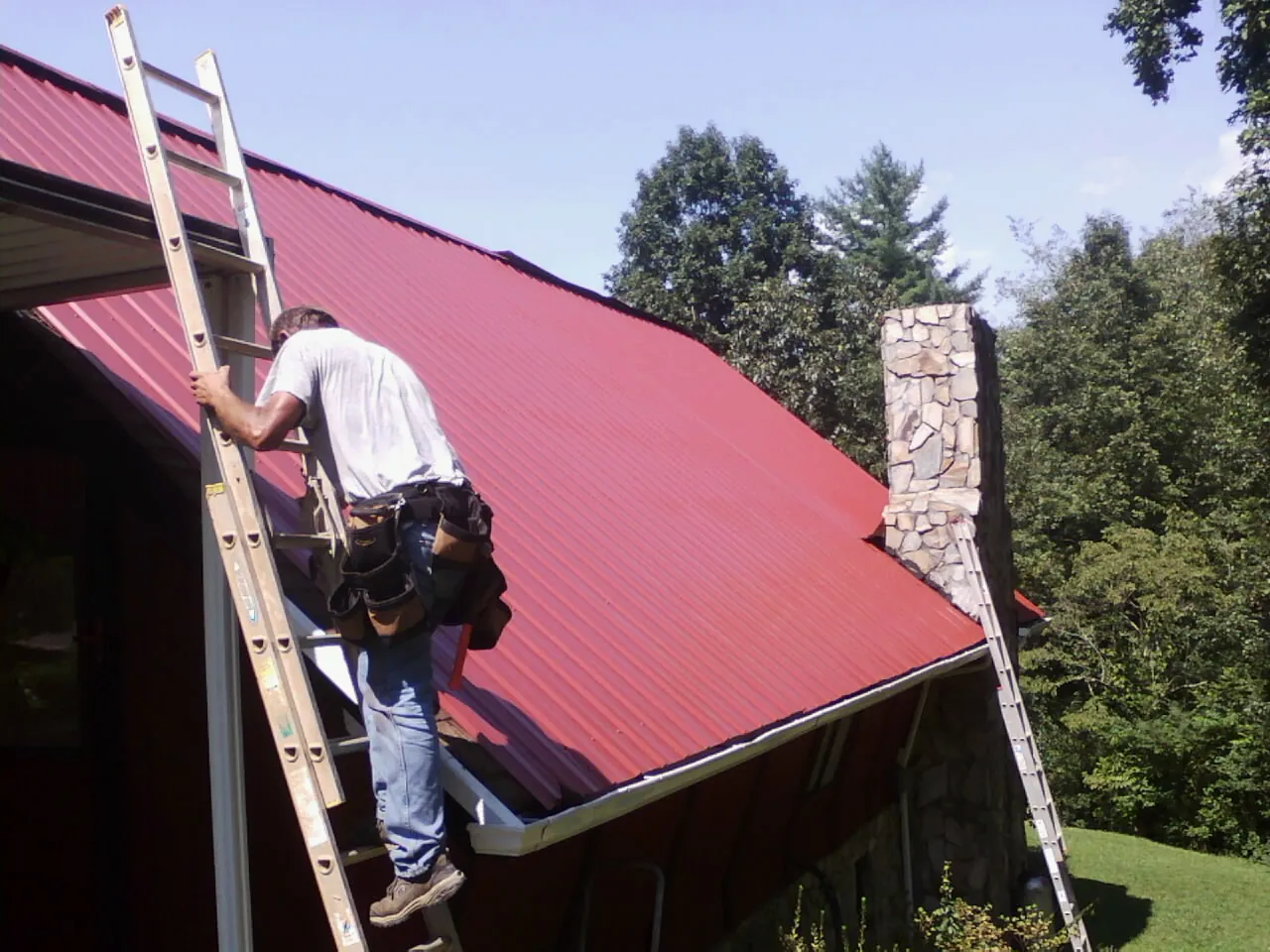 Roofer on a ladder beside a red metal roof and stone chimney — Soffit & Fascia Repair by Roofing Brothers in Low Moor